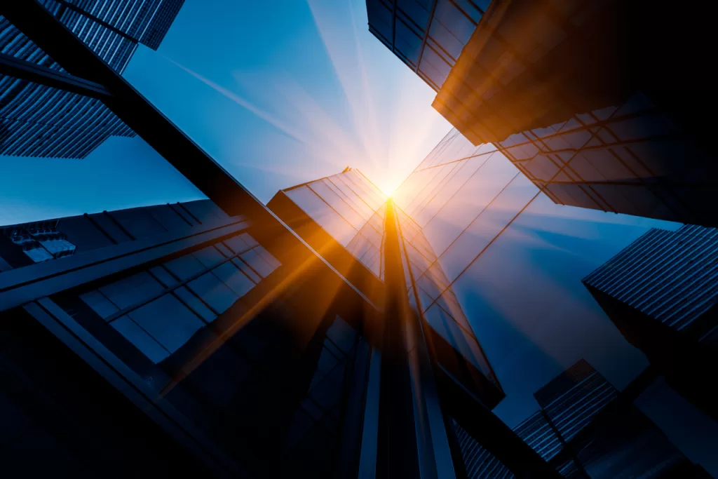 A striking low-angle view of several modern glass skyscrapers with the sun shining between them, creating a dynamic light reflection and a futuristic urban feel.