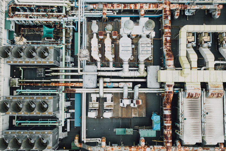 A high-angle photograph displaying an extensive rooftop HVAC system with ventilation ducts, cooling units, and pipes. Some parts of the infrastructure appear rusted, highlighting the impact of weathering on industrial systems.