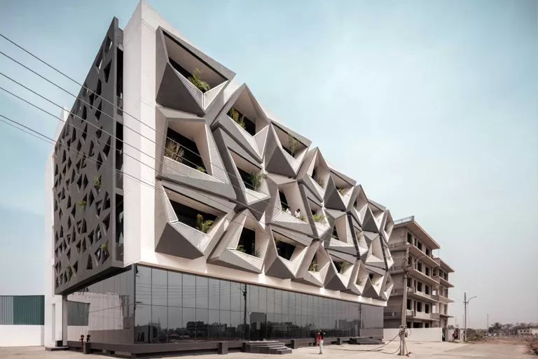A modern building with an intricate geometric façade featuring angular, protruding balconies filled with greenery. The lower part of the building has a reflective glass exterior, contrasting with the white and gray upper structure. The design incorporates both aesthetics and functionality, likely providing shade and ventilation. The background shows another building under construction in an urban setting.